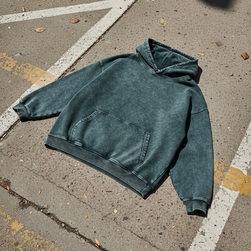 Garment-washed heavyweight hoodie in faded green, laid on asphalt showing the uneven wash across the seams